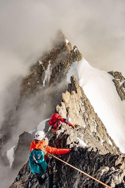Photo of two mountain climbers in orange on a mountain ridge. 